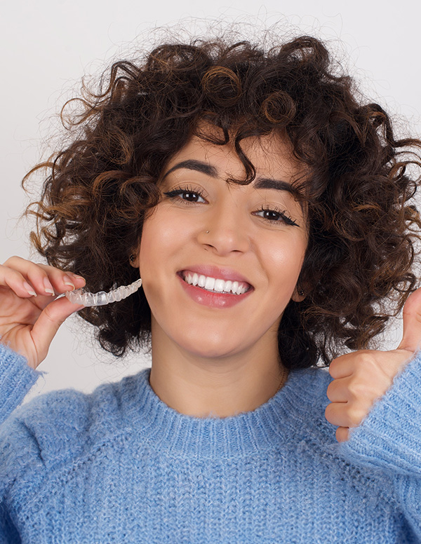 A woman with curly hair and a blue sweater is smiling at the camera, holding a toothbrush in her right hand.